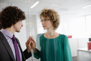 Deux collègues face à face au bureau derrière une vitre, illustrant les relations de couple au travail et les tensions possibles dans l’équipe.